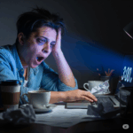 暗い部屋でノートパソコンに向かって作業している男性。顔は疲れていて、目の下にはクマがあり、口を大きく開けてあくびをしている。画面には午前3時と表示されており、使い捨てカップやクシャクシャになった紙が散らかった机の上には、作業用のランプが置かれている。