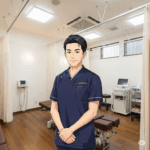 
笑顔で立つ若い男性の施術者が、胸の前で手を組み、カメラを見ているイラスト。背景には、施術用のベッドや医療機器が見える、明るく清潔感のある接骨院の室内が描かれている。