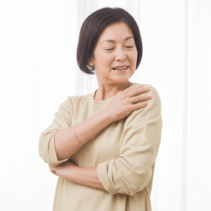 窓辺に立つ50代くらいの日本人女性が、自分の右肩に手を当てて微笑んでいる。ベージュの長袖シャツを着ている。背景は白いカーテン。