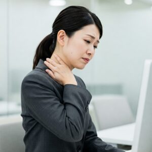 オフィスでパソコンに向かい、首に手を当てて辛そうな表情をしている40代の日本人女性。グレーのジャケットを着ている。背景は明るいオフィス環境。